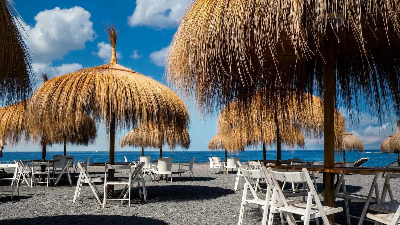 Serene beach scene with thatched umbrellas and chairs under a sunny sky in Adeje, Spain.